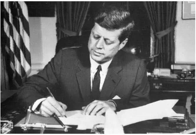 Fotografia in bianco e nero: il presidente Kennedy seduto alla scrivania, chino sulla carta, in atto di firmare.